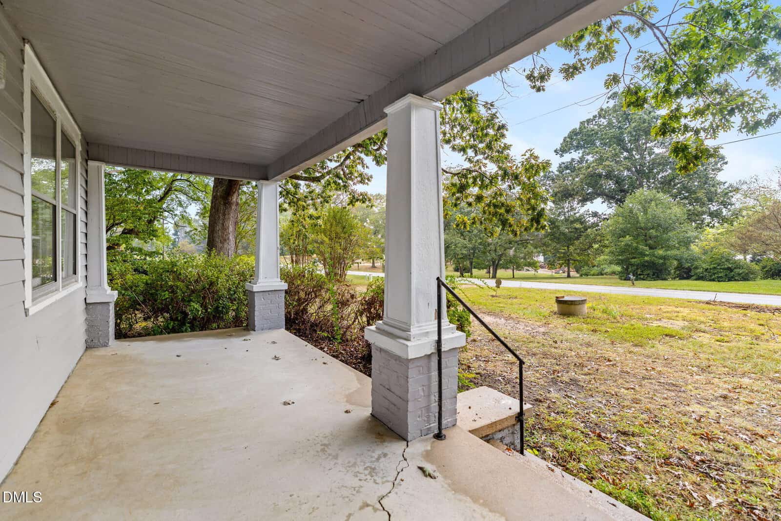 A covered concrete front porch with white pillars overlooks a grassy yard with bushes and trees. Steps with a metal railing lead down to the lawn, and a quiet street is visible in the background.