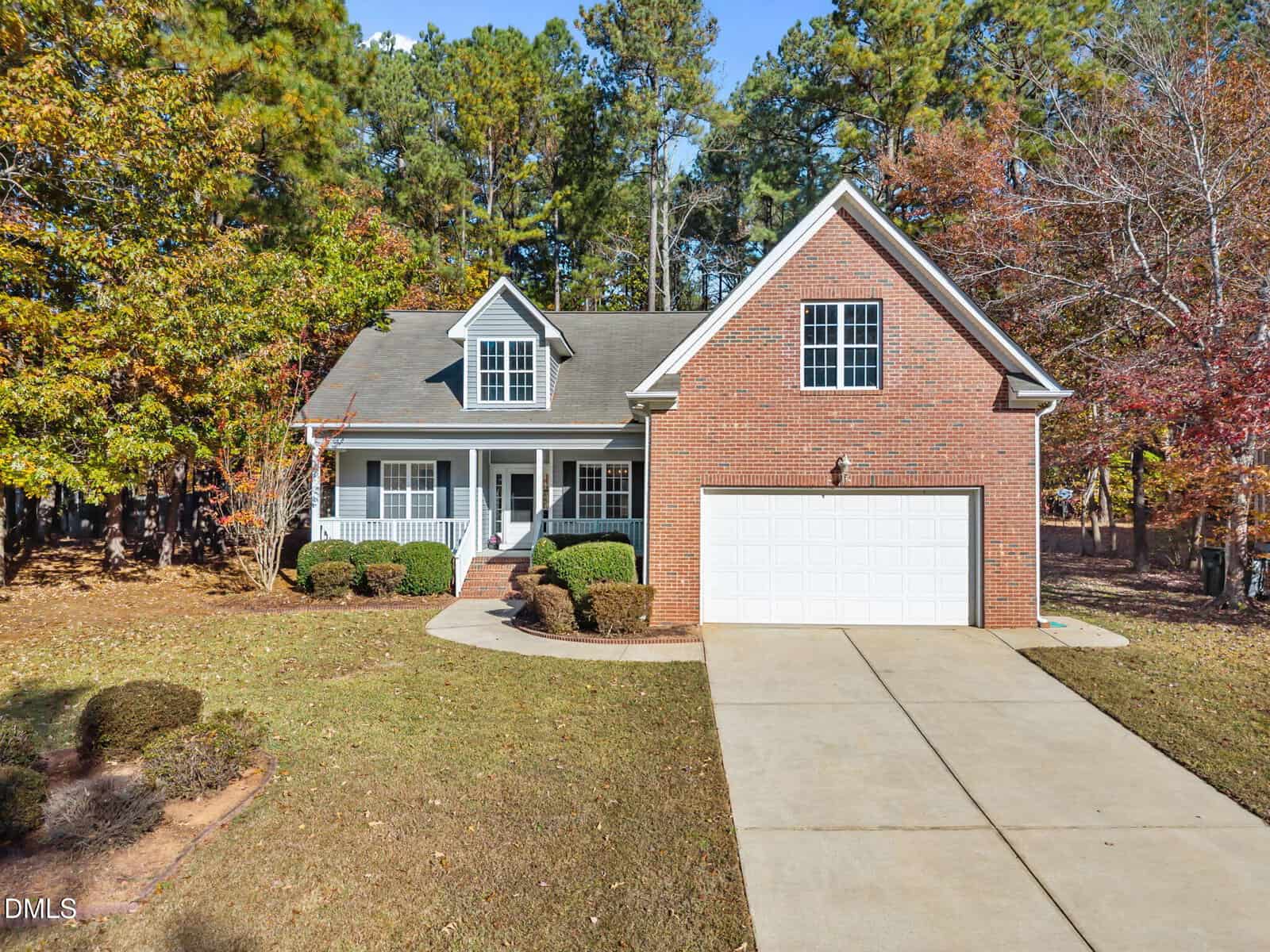 A brick house at 140 Applegate Drive, Franklinton, featuring a two-car garage, front porch, and dormer window, surrounded by trees and a lawn with trimmed bushes. A concrete driveway leads to the garage.