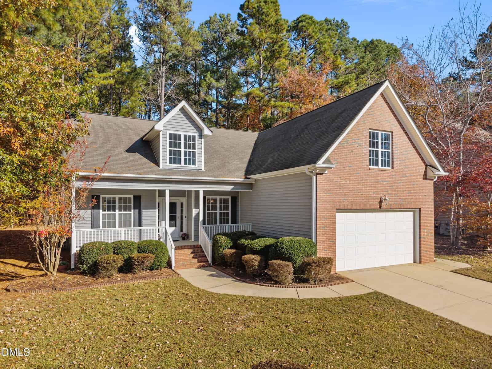 A two-story suburban house with gray siding, a brick section, white trim, a covered front porch, dormer windows, attached two-car garage, and manicured bushes, set against a backdrop of tall trees and a blue sky.