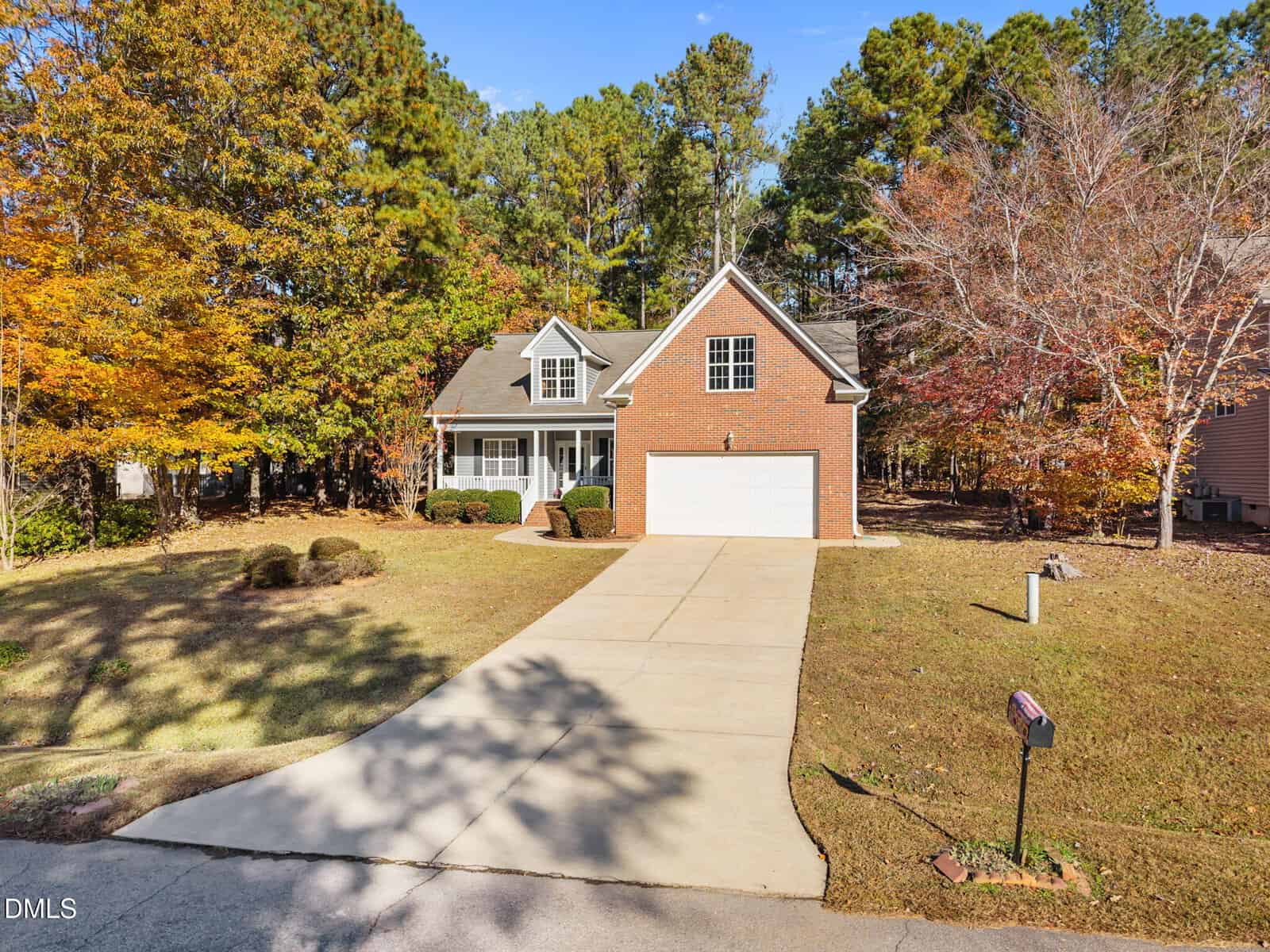 A two-story suburban house with brick and siding exterior, attached two-car garage, long driveway, and a front yard with minimal landscaping. Trees with autumn foliage surround the property.