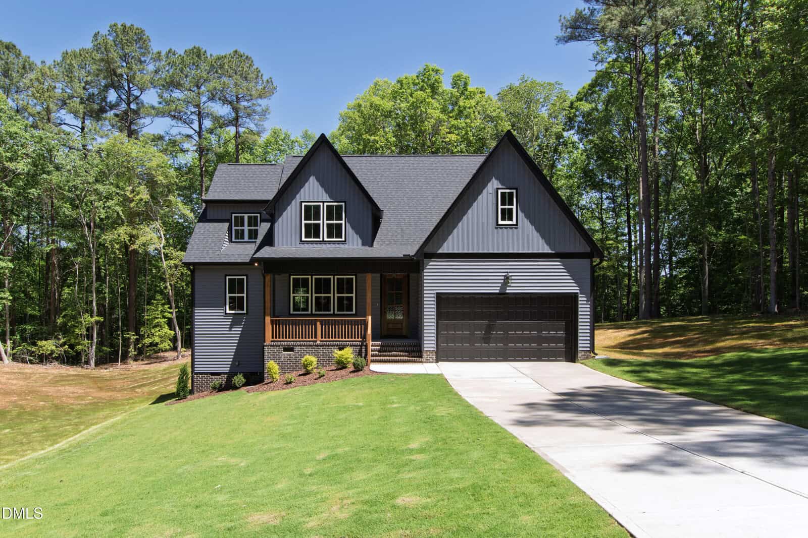 A modern gray house with black trim at 212 Rawhide Drive in Louisburg features a covered front porch and attached two-car garage, set on a well-manicured, sloping lawn surrounded by tall trees under a clear blue sky.