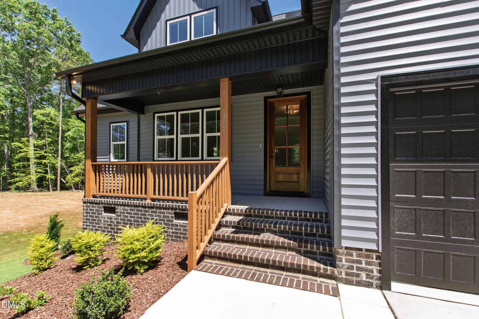 A modern house exterior with gray siding, black trim, a wooden front door, porch with wooden railings, brick steps, and landscaped shrubs beside a concrete walkway.