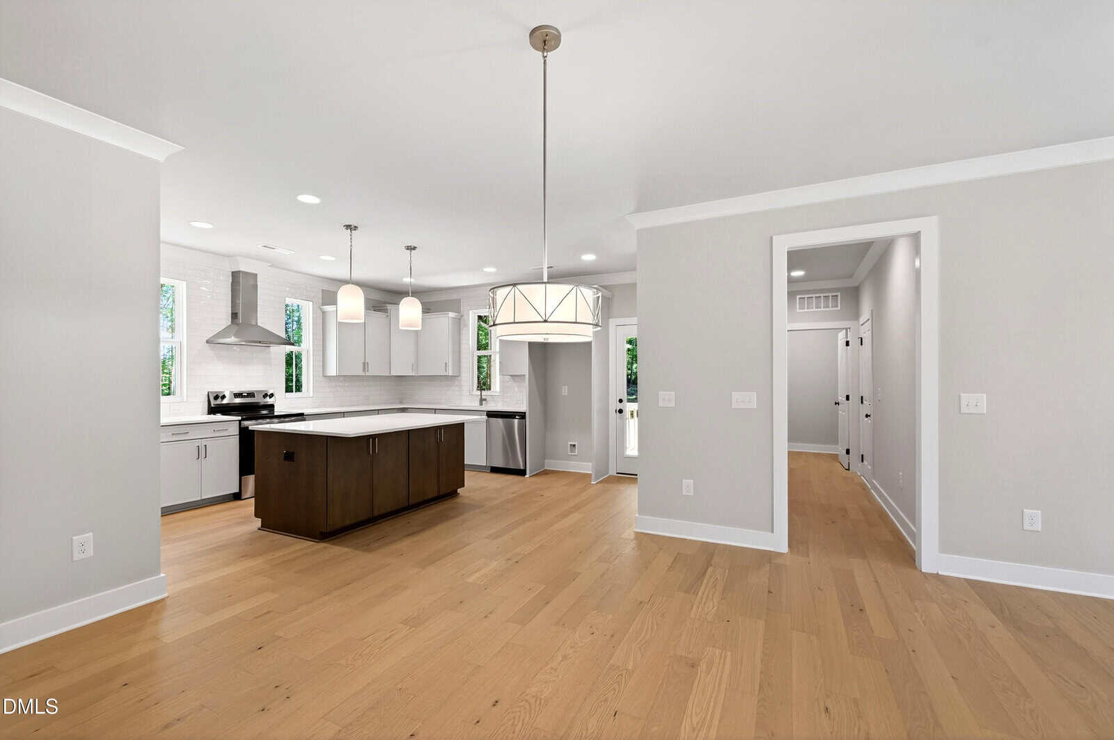 Modern open-concept kitchen and dining area with light wood floors, white walls, stainless steel appliances, pendant lighting, and an island with dark cabinetry. Multiple doorways lead to other rooms.