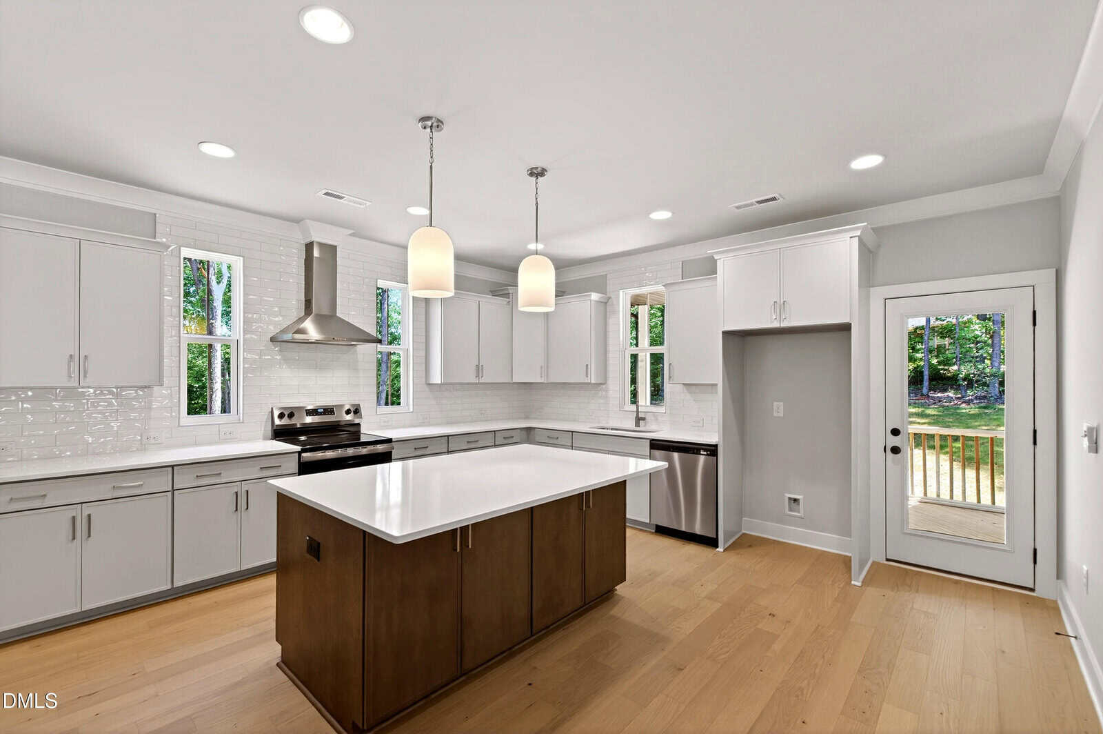 Modern kitchen with white cabinets, stainless steel appliances, a large island with a white countertop, wooden floors, pendant lighting, and windows providing natural light with a view of greenery outside.