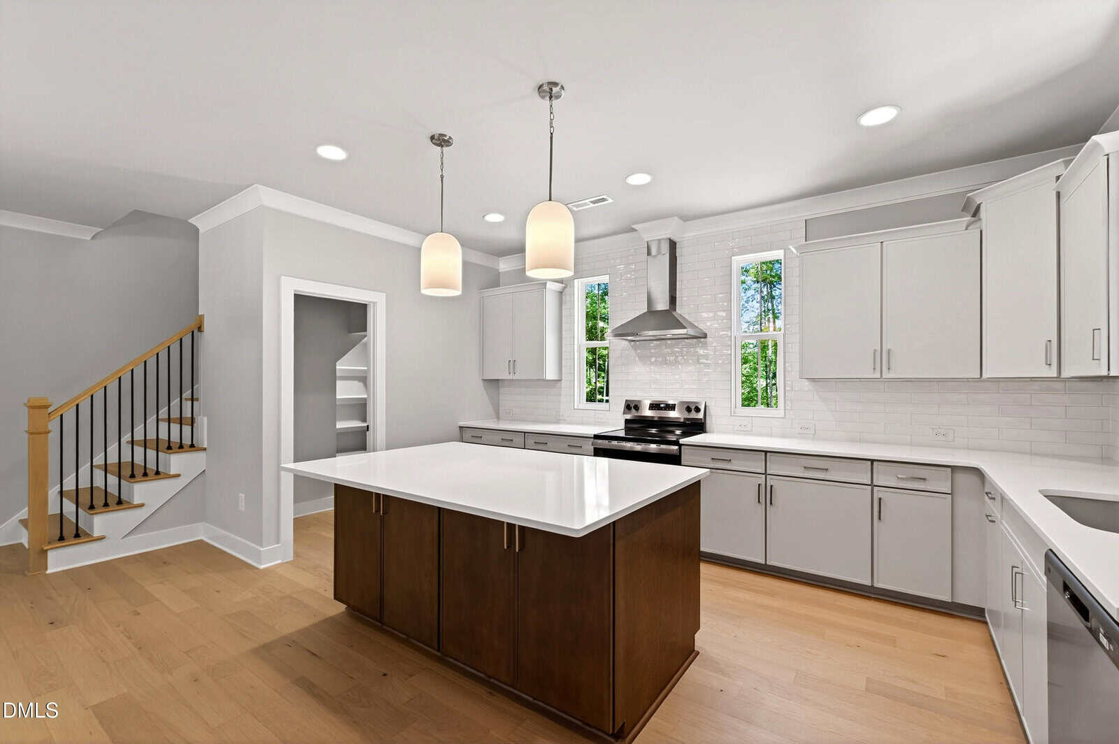 Modern kitchen with white cabinets, a center island with dark wood, stainless steel appliances, pendant lights, wood flooring, and two windows above the sink. A staircase and walk-in pantry are visible in the background.