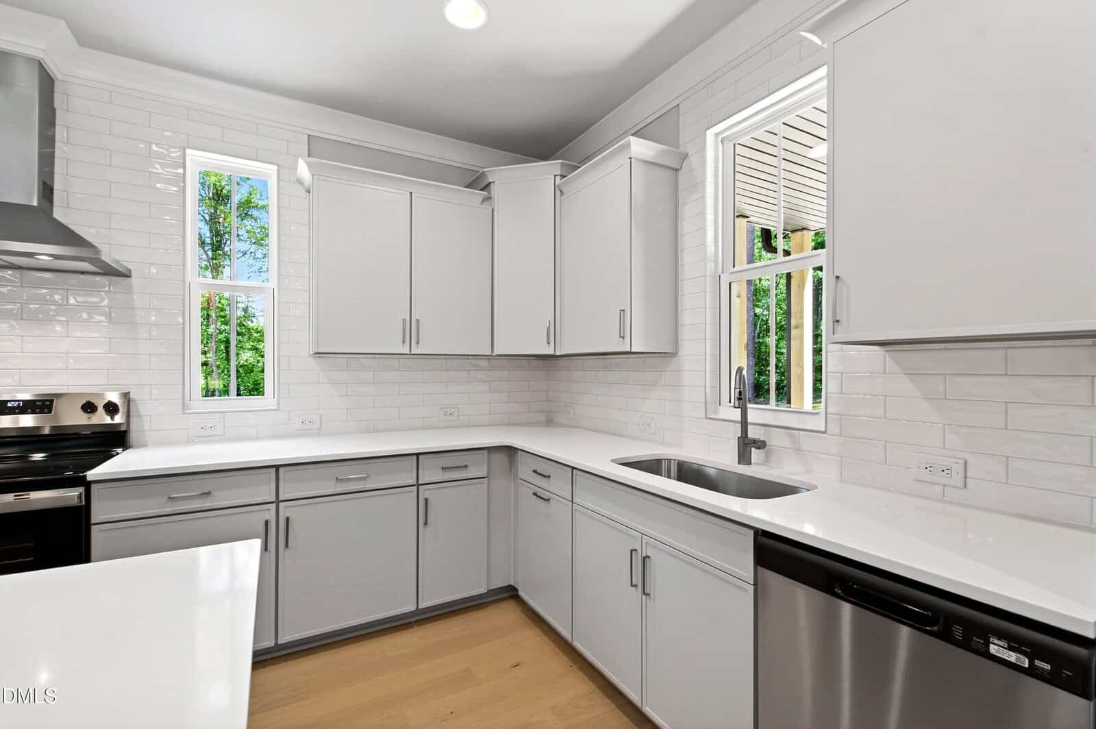 Modern kitchen with light gray cabinets, white countertops, stainless steel appliances, white subway tile backsplash, two windows with greenery outside, and light wood flooring.