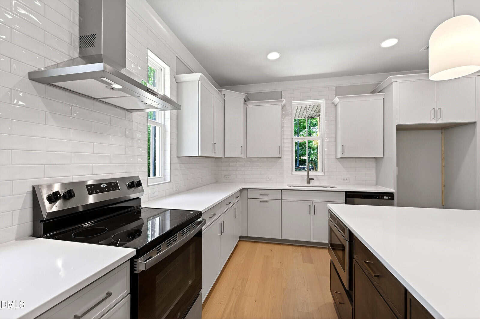 Modern kitchen with white cabinets, stainless steel stovetop and range hood, center island with dark wood base, light wood flooring, white subway tile backsplash, and two windows above the sink letting in natural light.