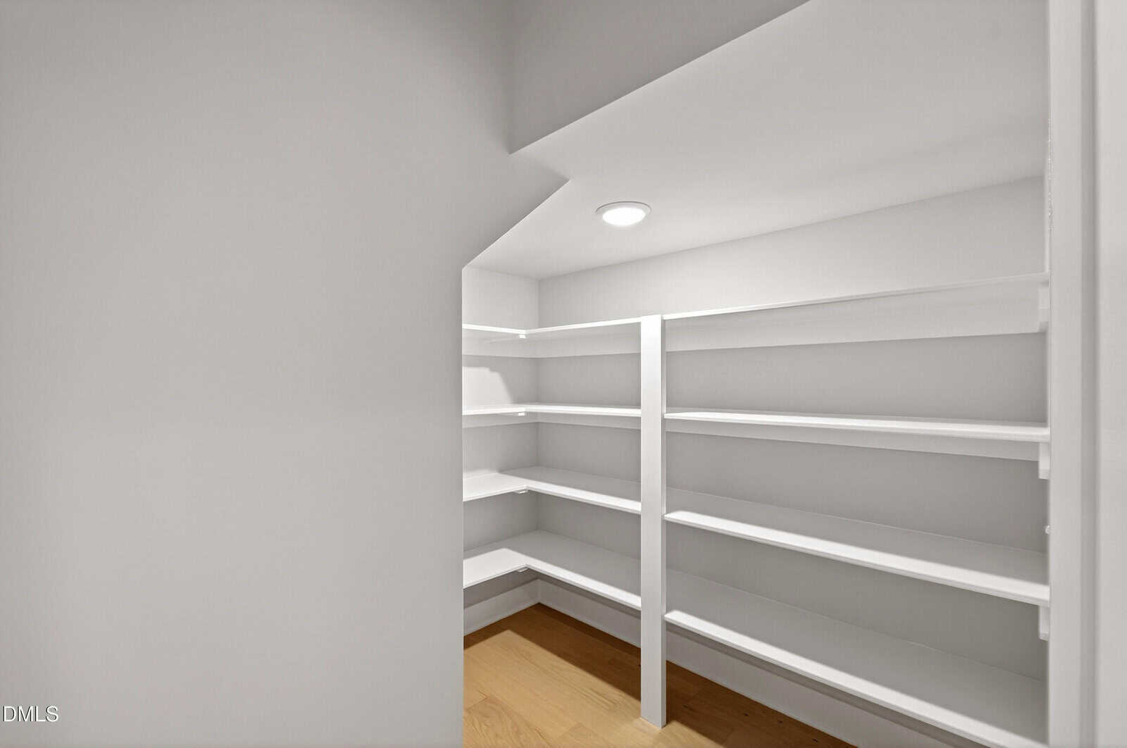 A small, white walk-in pantry with angled ceiling, white shelves on two walls, and a light wood floor. A ceiling light fixture provides illumination.