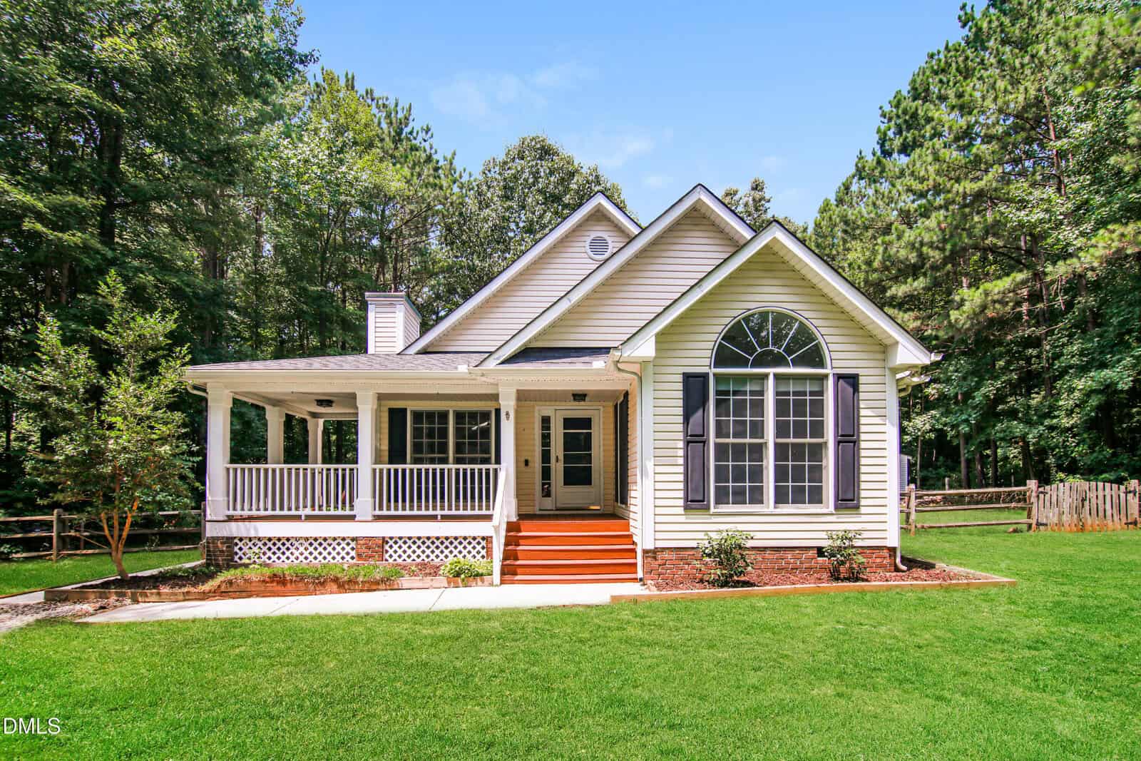 A single-story house with white siding, a front porch, large arched window, and black shutters stands on a green lawn surrounded by trees on a sunny day at 190 Hardwood Drive in Franklinton.