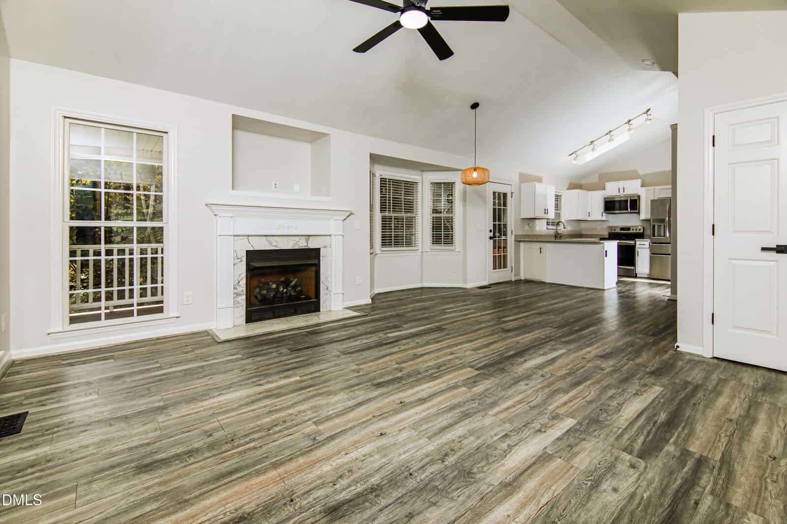 Spacious open-concept living area with wood-look flooring, a white fireplace, large windows, ceiling fan, and view into a modern white kitchen with stainless steel appliances and recessed lighting.