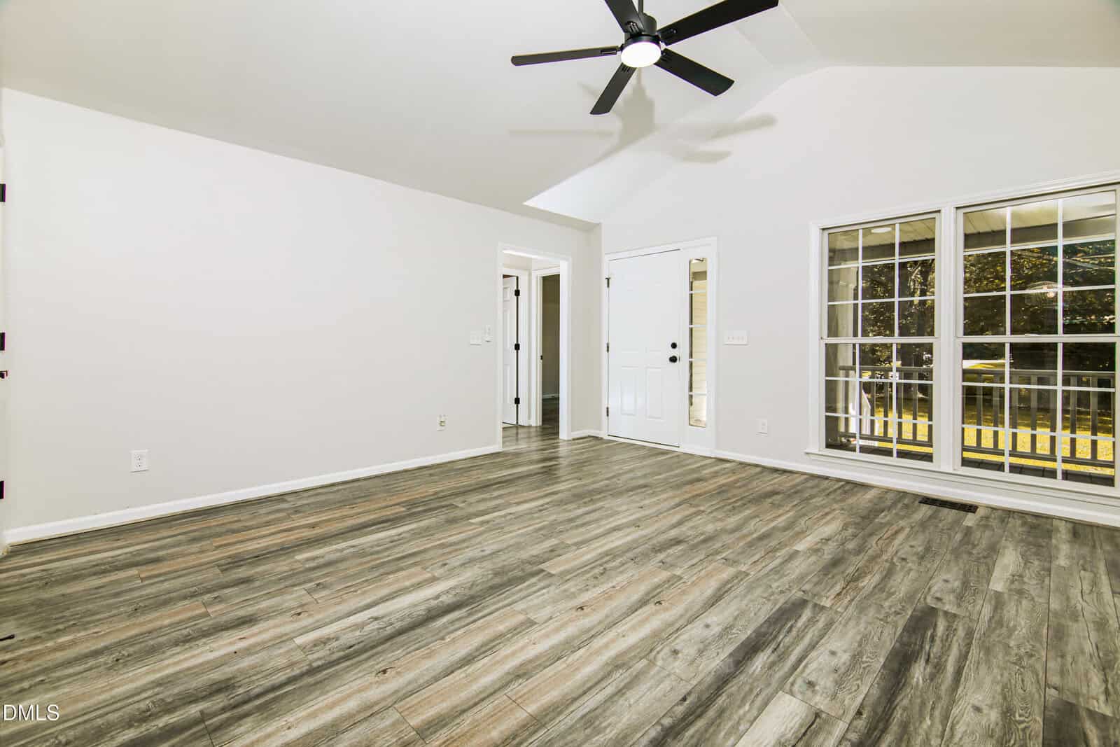 Empty room with wood-style flooring, white walls, a ceiling fan with lights, a door with sidelights, and large windows showing a view of a fenced yard with trees outside. The space is brightly lit and unfurnished.