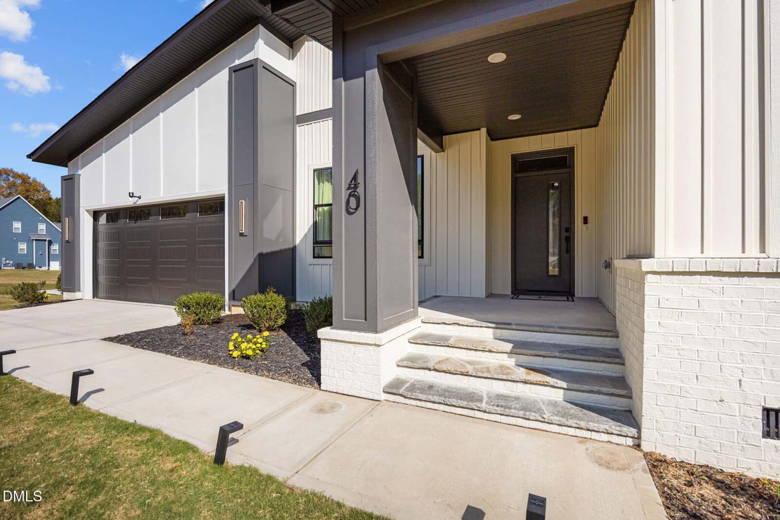 Modern house exterior with black and white siding, a two-car garage, covered front porch, black door, and steps leading to the entrance. The yard features green grass, shrubs, and outdoor lighting.