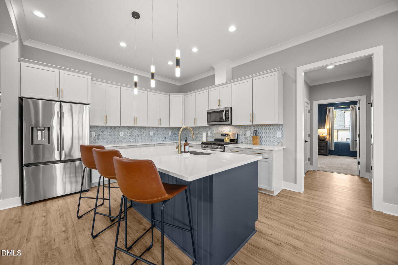 Modern kitchen with white cabinets, stainless steel appliances, a navy island with white countertop, three brown leather barstools, wood flooring, pendant lights, and a patterned backsplash. A hallway leads to another room.