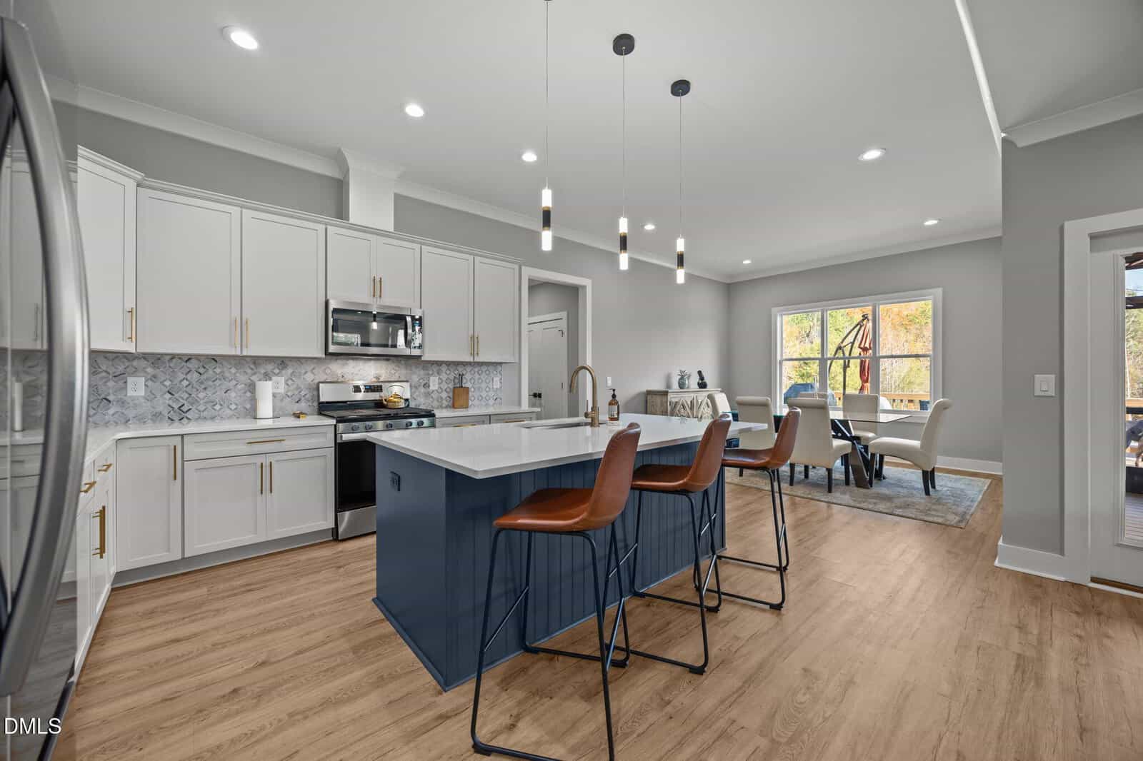 Modern kitchen with white cabinets, stainless steel appliances, a blue island with sink, three brown barstools, pendant lights, wood flooring, and an adjacent dining area with large windows.