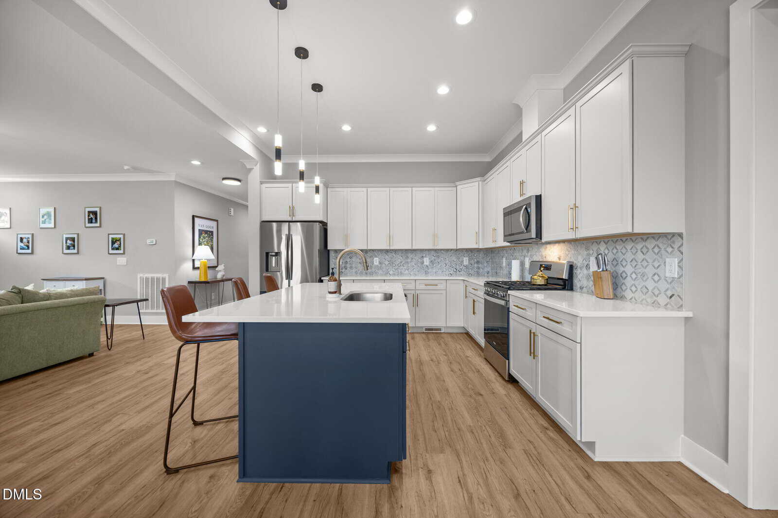 Modern kitchen with white cabinets, stainless steel appliances, a blue island with a sink, pendant lights, wood flooring, and barstools; open layout leads to living area with a green sofa and framed pictures on the wall.