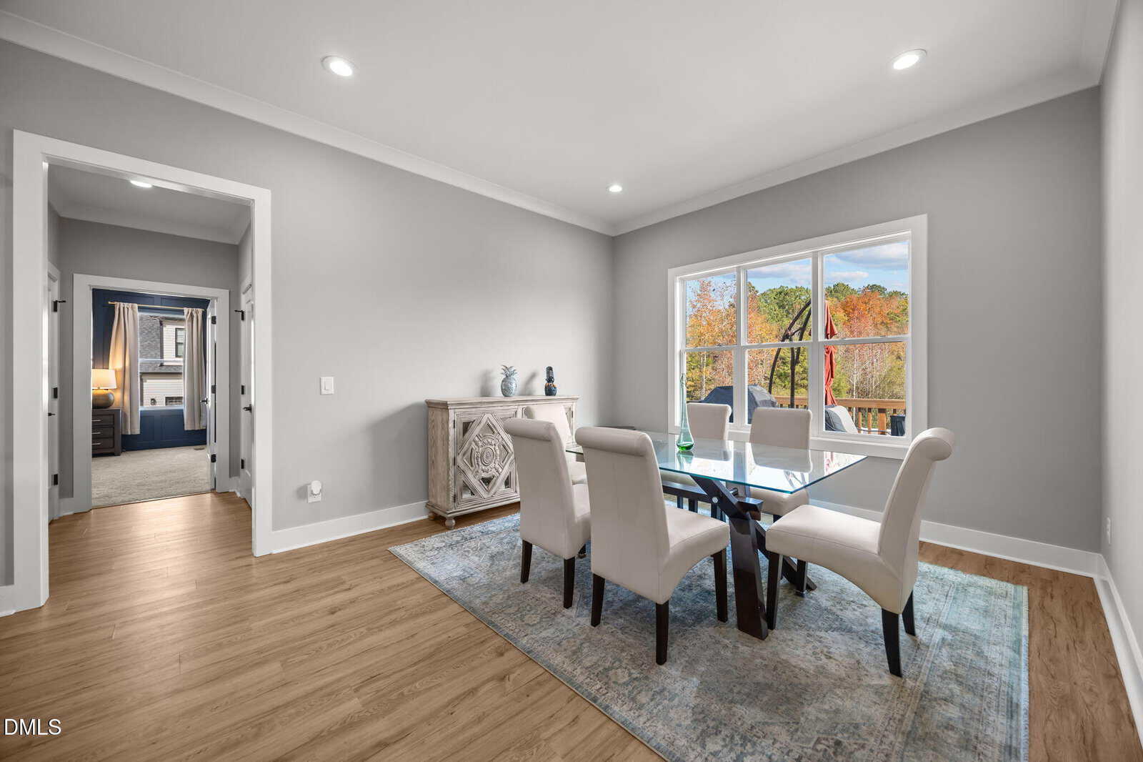 Bright dining room with a glass table, six white chairs, a decorative sideboard, wood flooring, large window showing trees outside, and a doorway leading to another room.