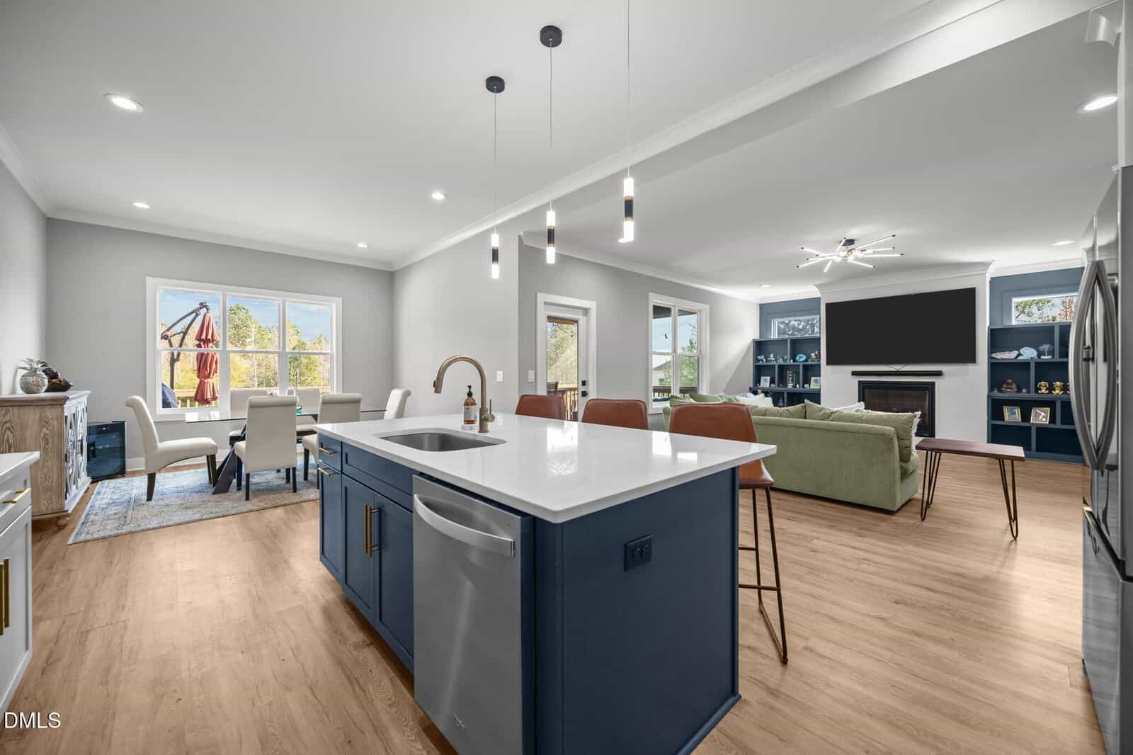Modern open-concept kitchen and living area with a blue island, bar stools, dining table, green sofa, wall-mounted TV, large windows, and wood flooring. Natural light fills the space.