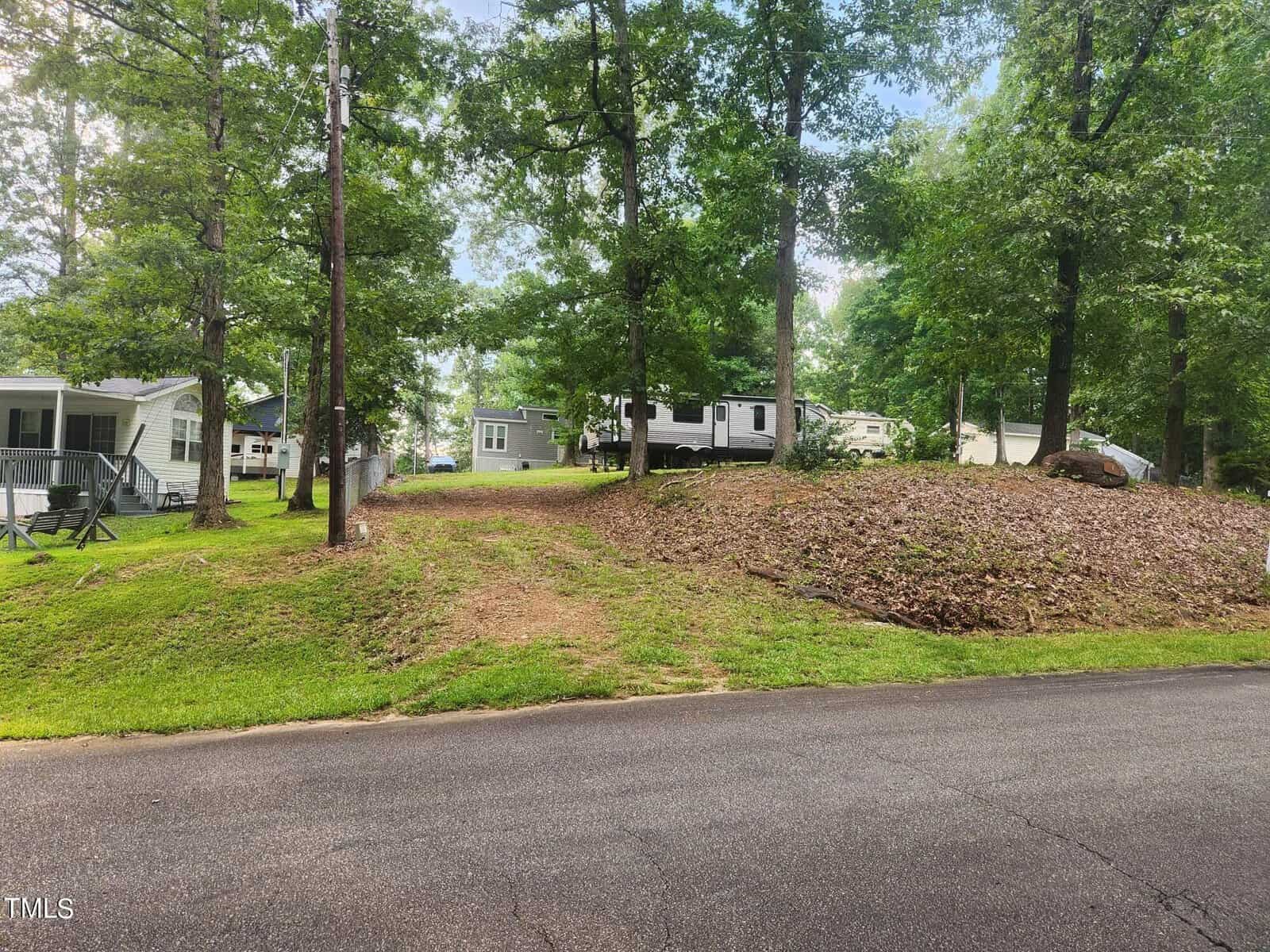 A paved road runs in front of two mobile homes and a white RV set among tall trees. A dirt driveway leads up a small hill to the homes, and the area is surrounded by green grass and scattered leaves.