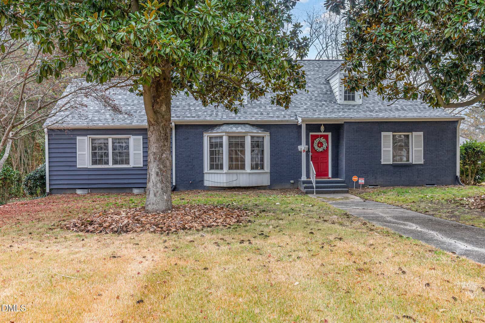 A single-story gray brick house at 612 N Main Street, Louisburg, features white shutters and trim, a red front door adorned with a wreath, multiple windows, and a large tree in the front yard with a patchy lawn and scattered leaves.
