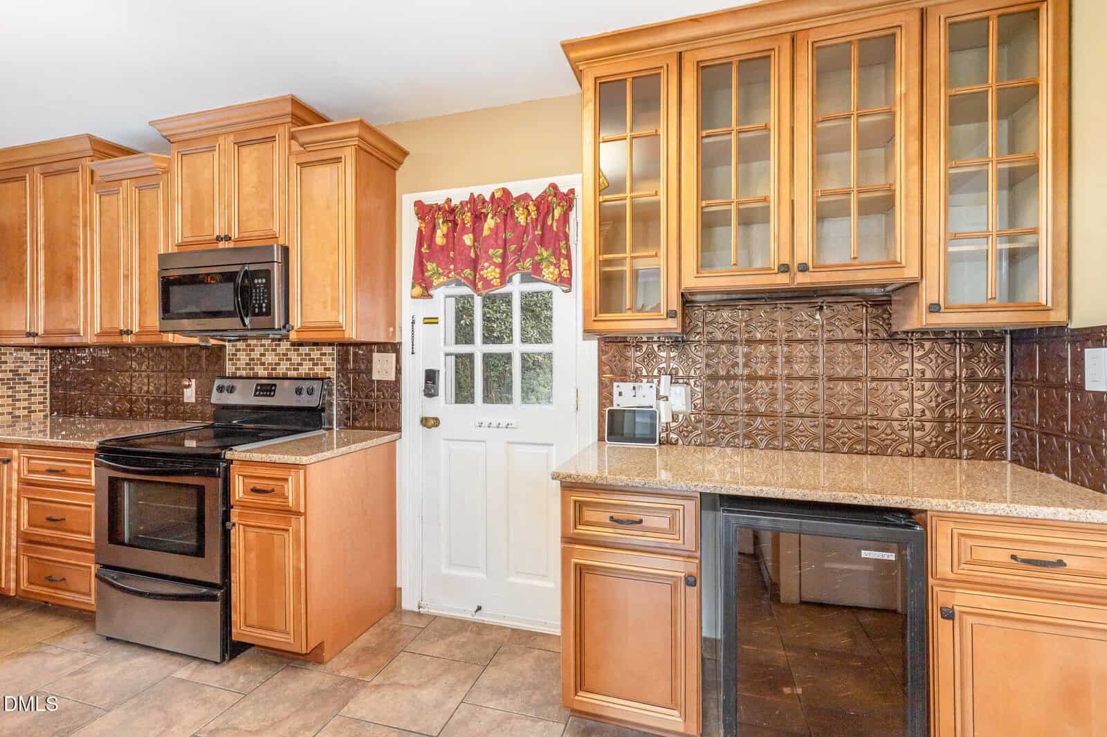 Kitchen with wood cabinets, stainless steel oven and stove, brown tile backsplash, granite countertops, glass-front cabinets, small wine fridge, and a white door with a window and red floral valance. Brown tile floor is visible.