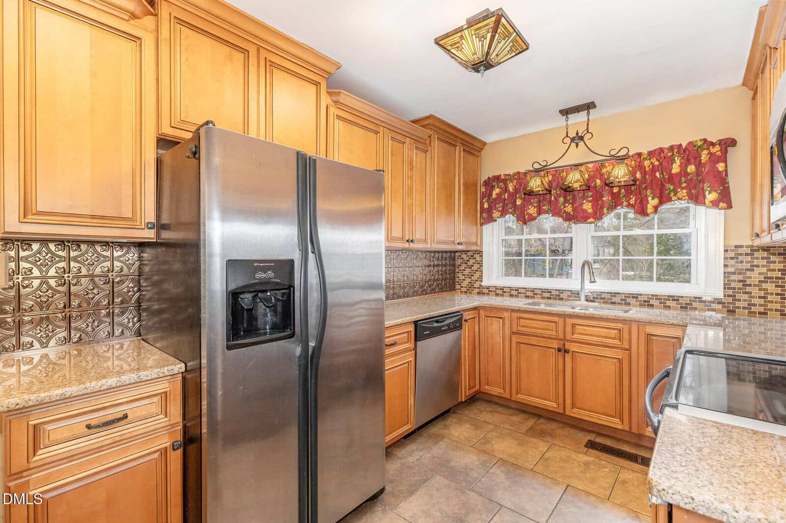 Kitchen with wooden cabinets, stainless steel appliances including a refrigerator, dishwasher, and oven, beige granite countertops, a tile backsplash, and a double window above the sink with a red patterned valance.
