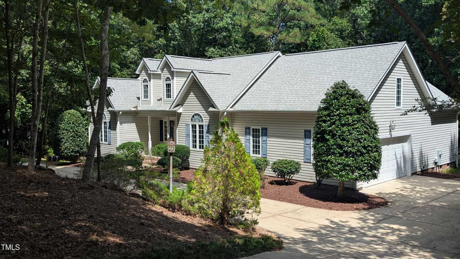 A beige single-story house with white trim and a gray shingle roof, surrounded by trees and shrubs, awaits you at 270 Lumberjack Lane in Louisburg. There is a driveway leading to a two-car garage and a small walkway to the front entrance.