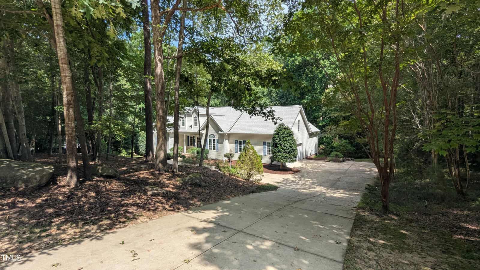 A white house with a light gray roof is partially visible behind trees. The house has arched windows and sits at the end of a long concrete driveway surrounded by wooded greenery on both sides.
