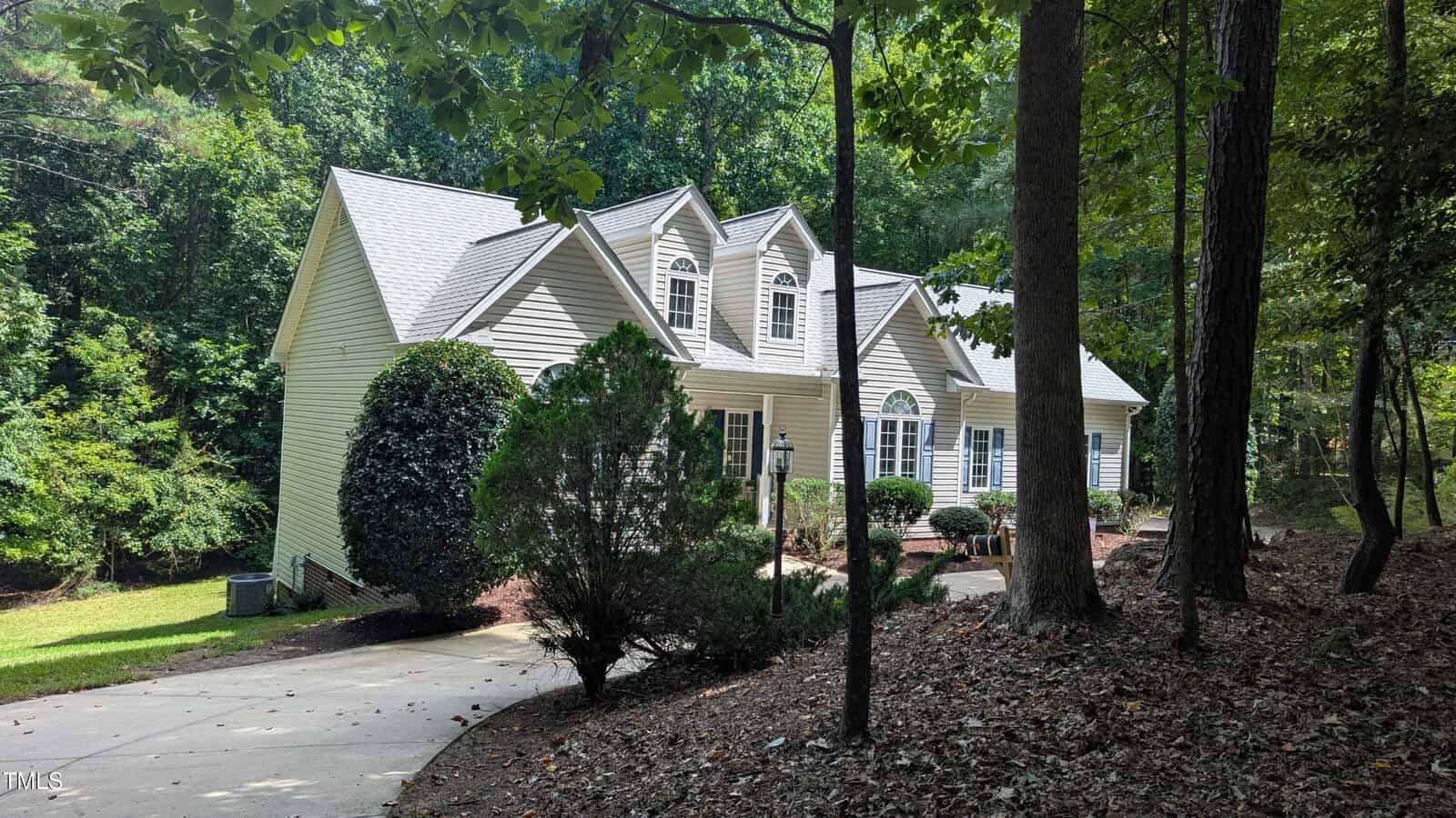 A light-colored, two-story house with dormer windows and a steep roof sits beside a curving driveway, surrounded by trees and shrubs in a wooded area.