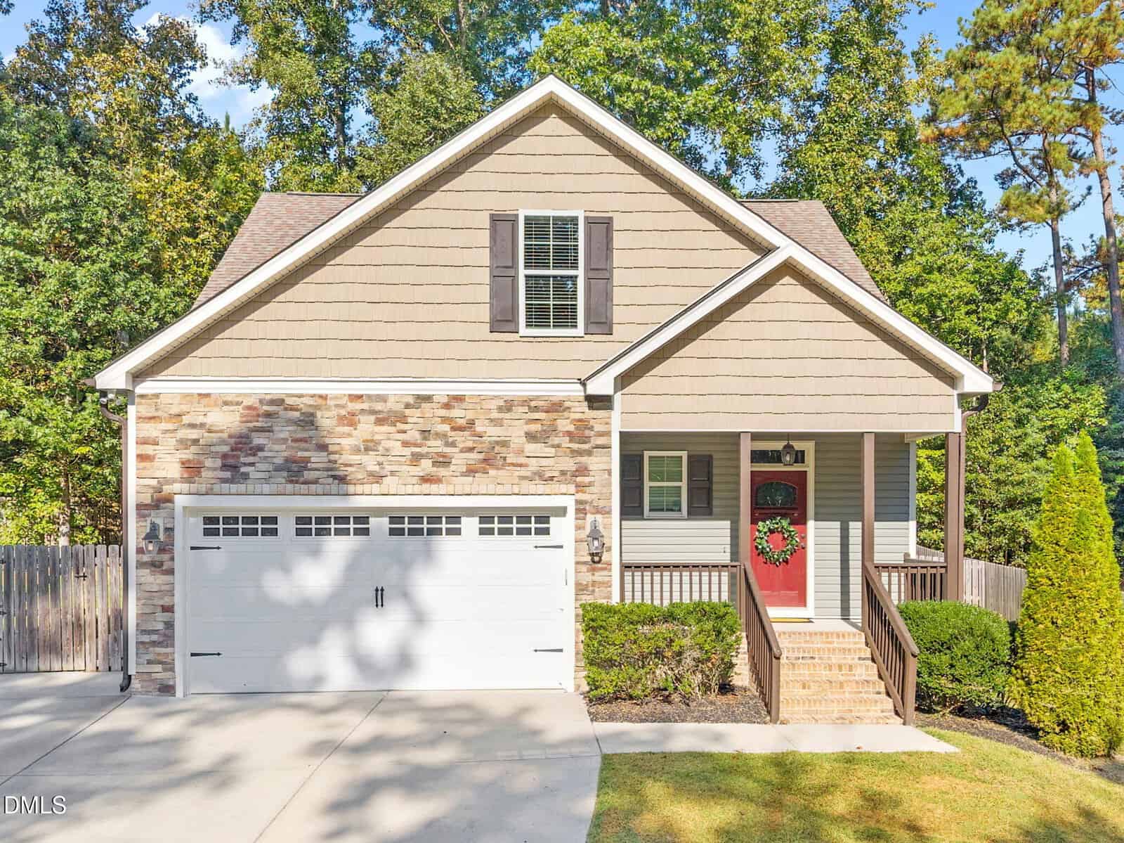 A two-story suburban house with tan siding, stone accents, a double garage, and a front porch with a red wreath on the door, surrounded by trees and greenery.