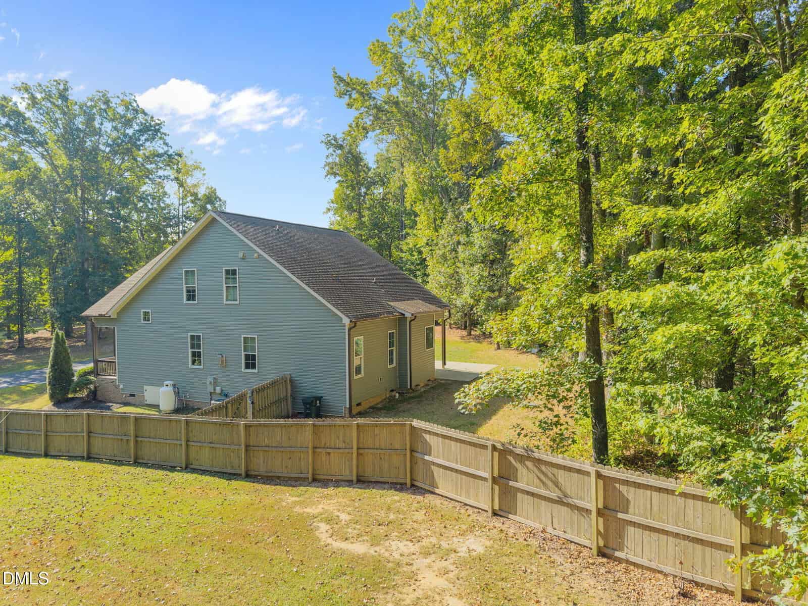 A light gray two-story house with a sloped roof sits beside a wooden fence and surrounded by tall, leafy trees under a clear blue sky, with a small backyard area visible.