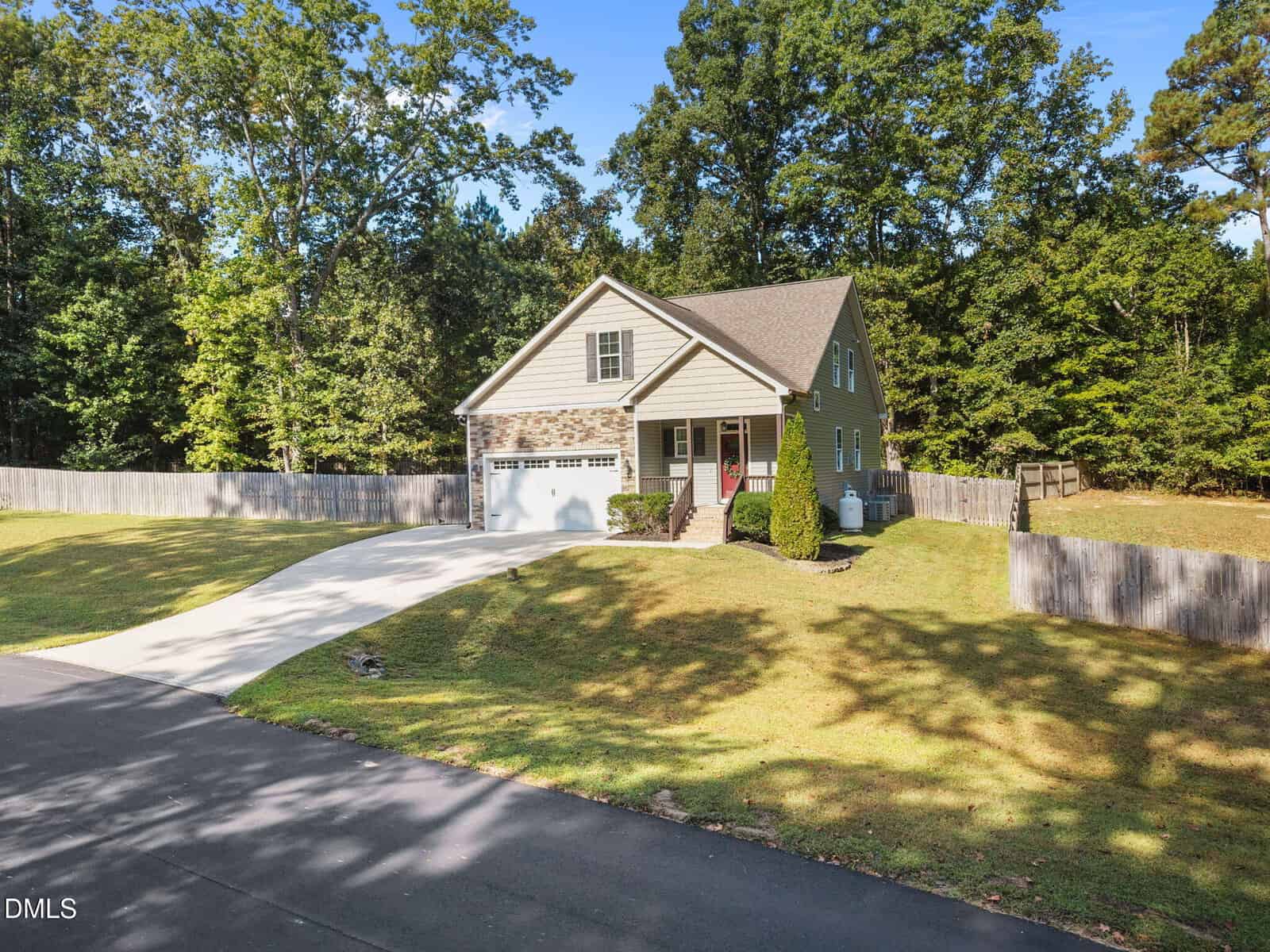 A two-story beige house with a gable roof, attached two-car garage, and a neatly mowed lawn, surrounded by a wooden fence and trees, with a curved driveway leading to the road.