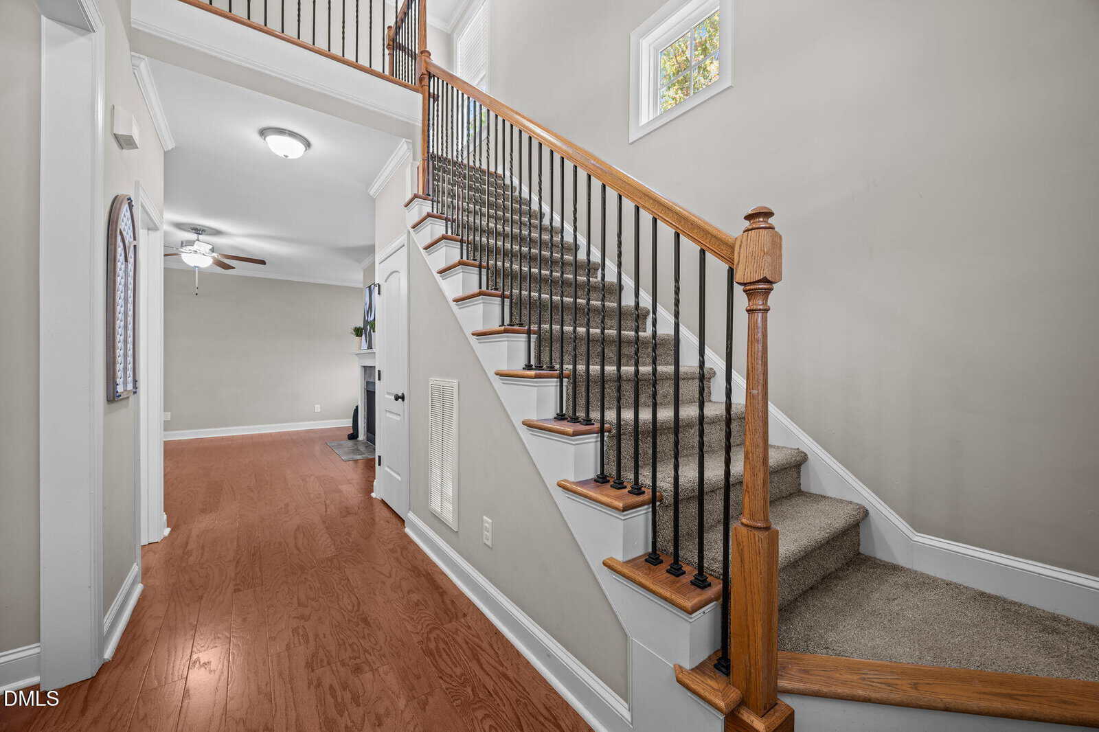 A carpeted staircase with wooden handrails and black balusters ascends from a hallway with wood flooring. The walls are light-colored and a ceiling fan is visible in an adjoining room. A small window lets in natural light.