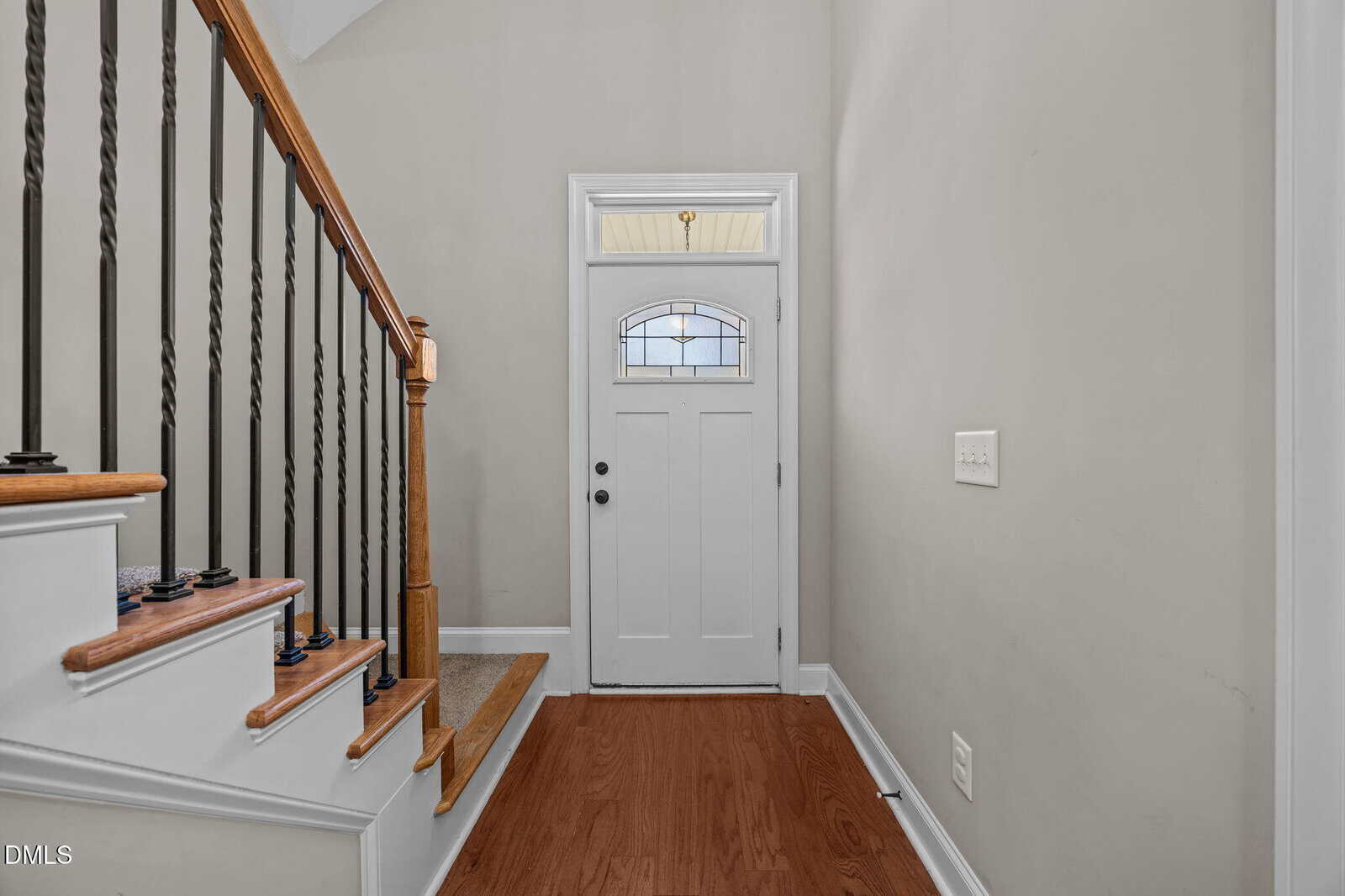 A white front door with a decorative window is set in a beige entryway. To the left, a wooden staircase with black iron railings leads upstairs. The floor is medium-toned wood, and the walls are light-colored.