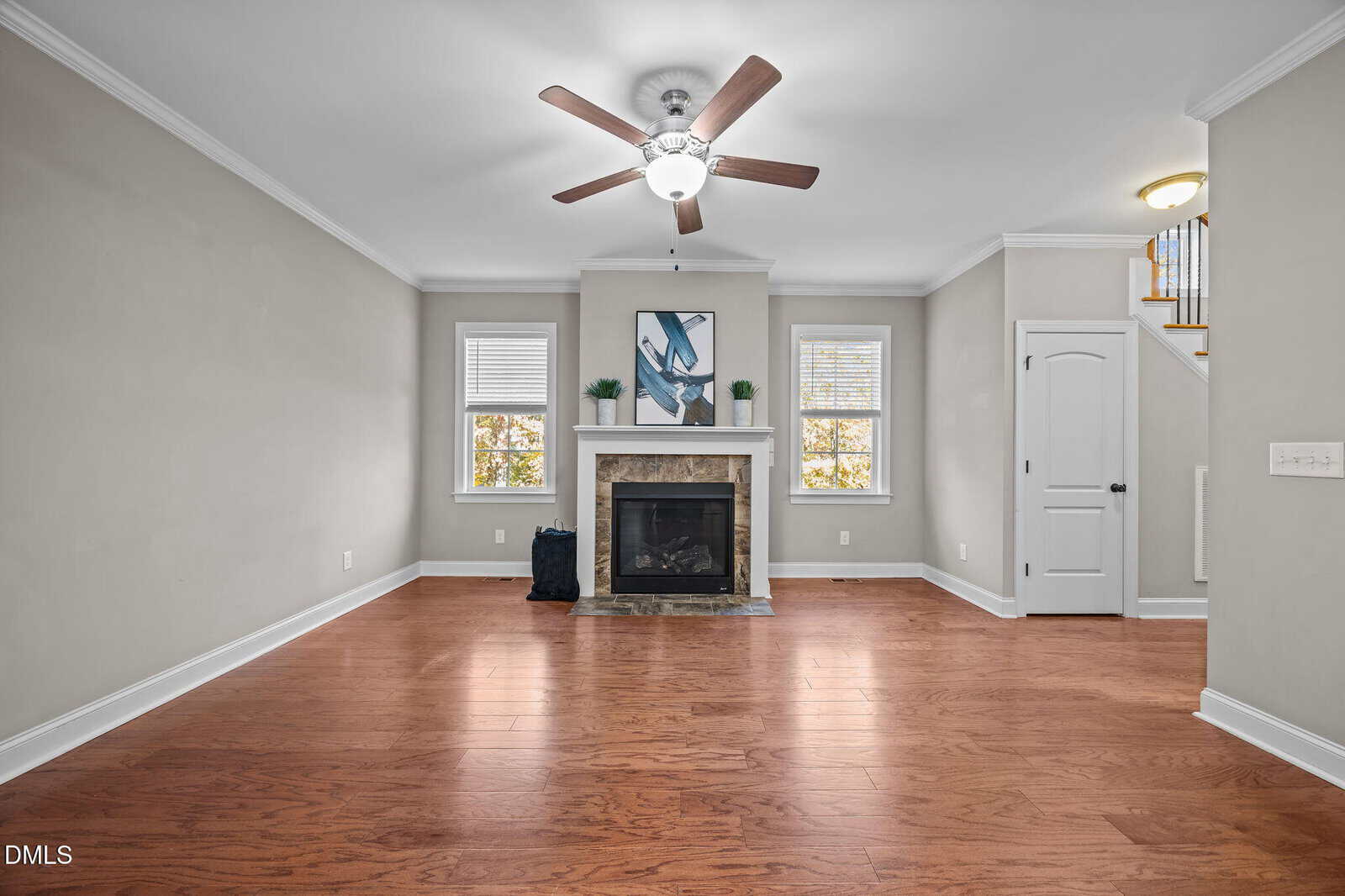 Bright living room with hardwood floors, a ceiling fan, fireplace with art above it, two windows on either side, and a door to the right leading to a staircase. Walls are painted light gray.