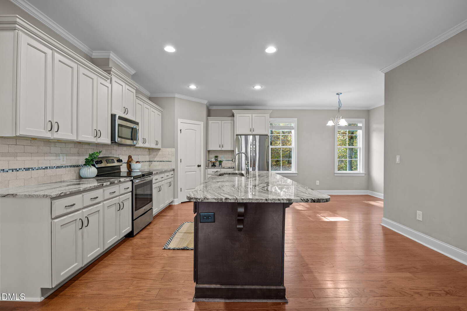 Modern kitchen with white cabinets, stainless steel appliances, a large marble island with a sink, and hardwood floors. There are two windows in a dining nook, letting in natural light. The walls are painted light gray.
