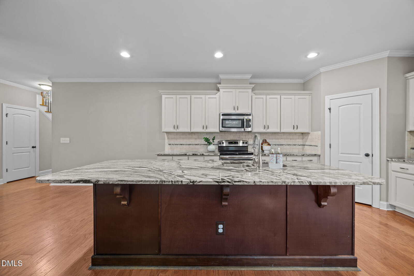 Spacious modern kitchen with a large marble island, stainless steel appliances, white cabinets, and wood flooring. Recessed lighting brightens the room, and a white door and stairway are visible in the background.