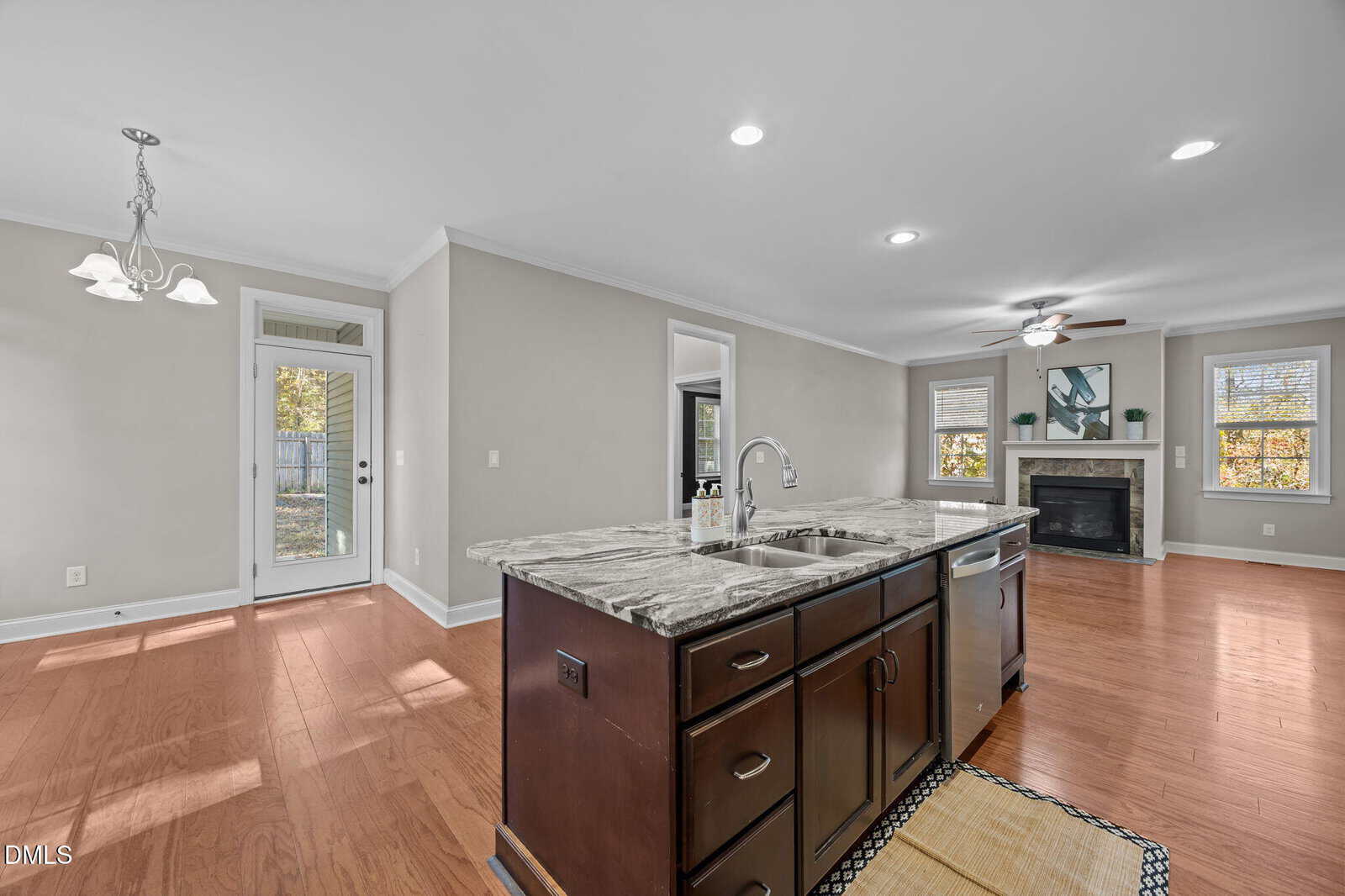 Open-concept kitchen and living area with wood floors, a marble island with sink, modern light fixture, fireplace, large windows, and a glass door leading to a backyard.