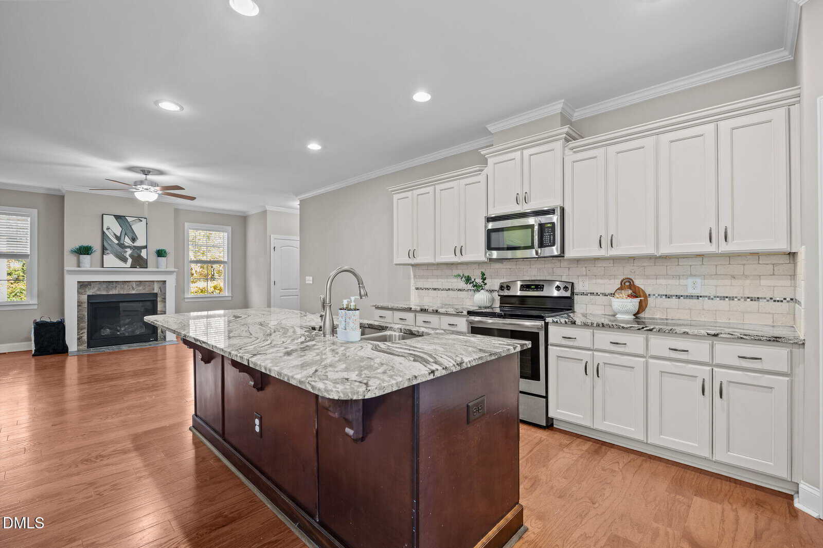 Modern kitchen with white cabinets, stainless steel appliances, a marble island with sink, wooden floors, and an open view to a living area with a fireplace and large window.