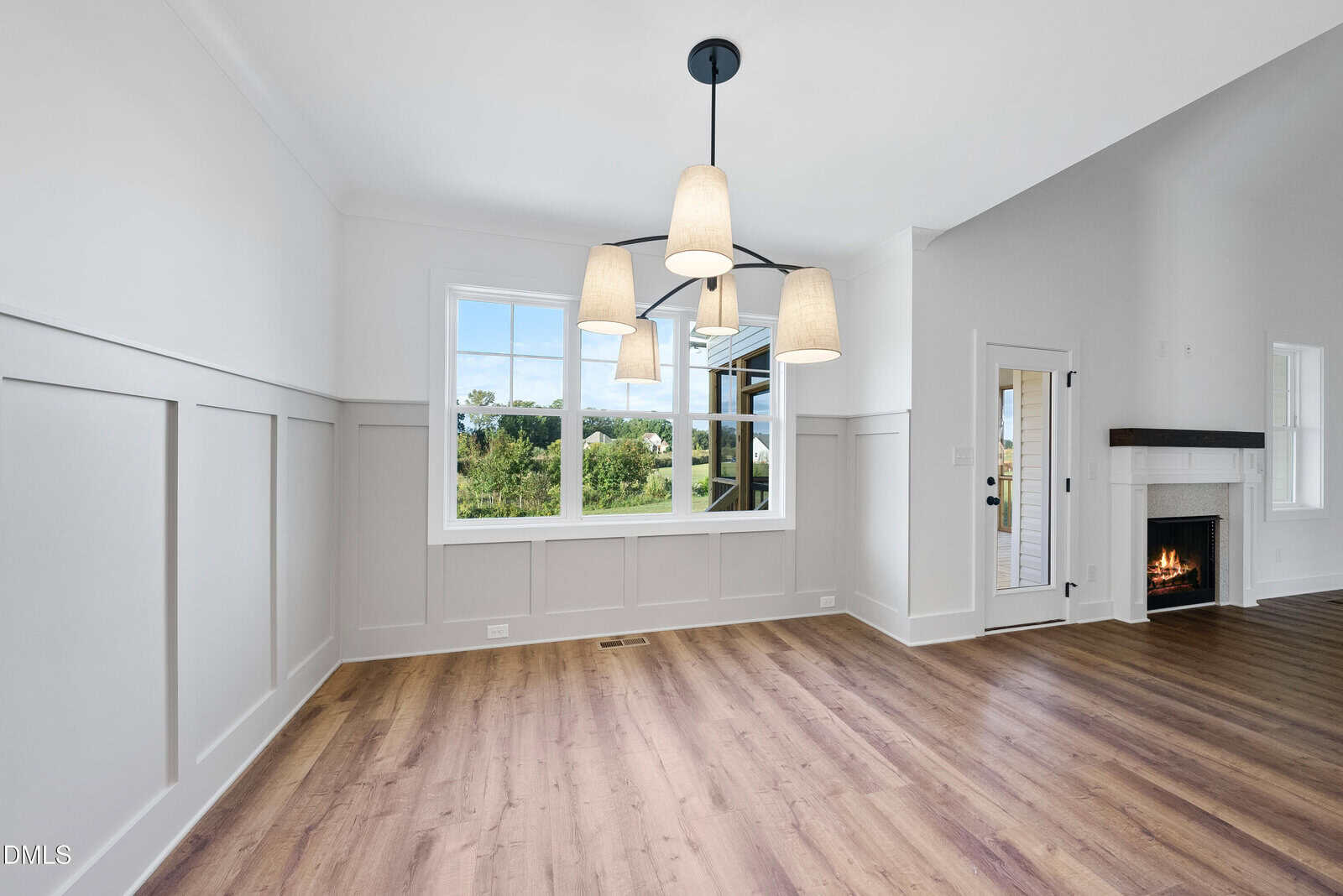 A bright, empty room with white paneled walls, large window, wood flooring, and a modern chandelier. A fireplace with a wooden mantel is visible in the adjacent living area.