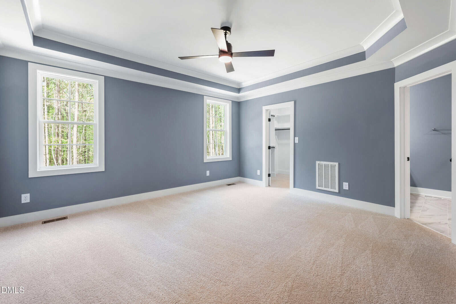 A spacious, empty bedroom with gray-blue walls, beige carpet, two large windows, a ceiling fan, and an open doorway leading to a tiled room. Natural light comes in from the windows.