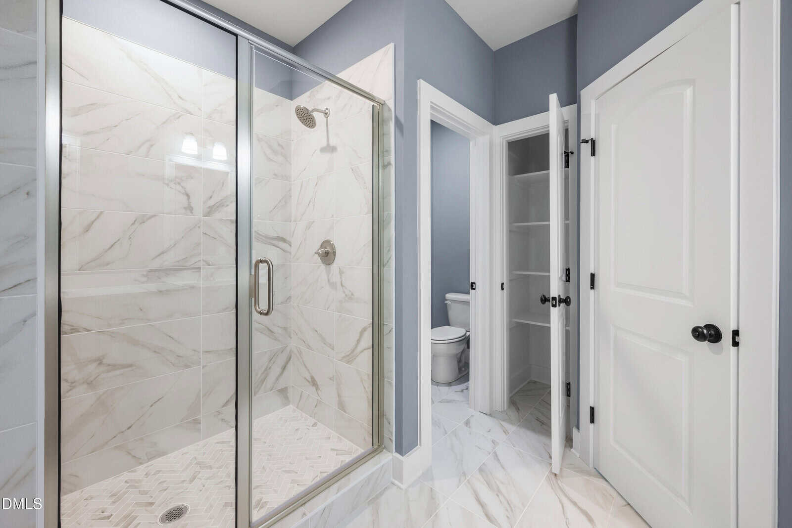 Modern bathroom with gray walls, a glass shower with marble tile, a white door closet with shelves, and a separate room with a toilet. The floor features large white marble tiles. Lighting fixture above the mirror.