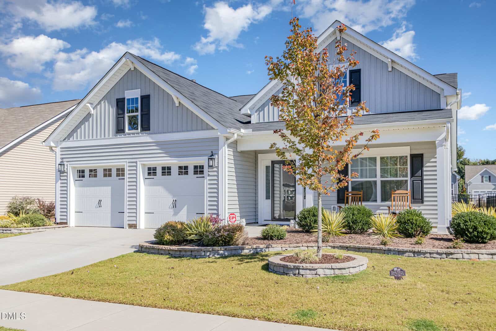 A modern two-story house with gray siding, white trim, and two garage doors. There is a small front porch with two chairs, a manicured lawn, landscaping, and a young tree in the yard under a blue sky with clouds.
