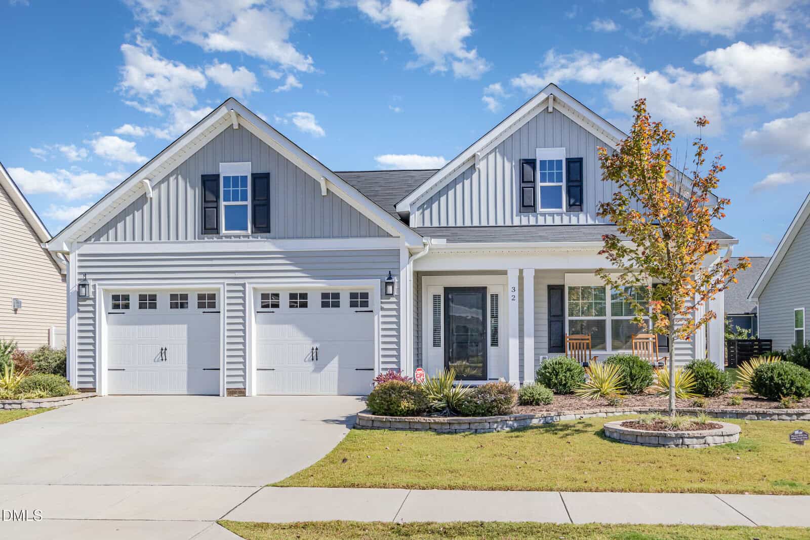 A modern gray house with white trim, two garage doors, a covered front porch with two chairs, and a landscaped yard with a small tree and shrubs under a partly cloudy sky.