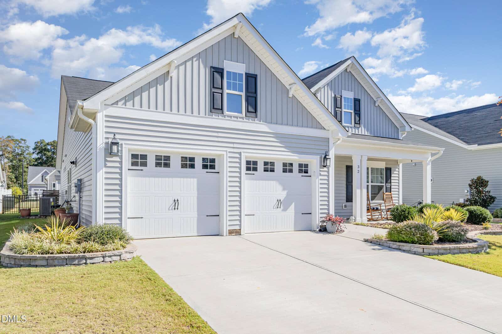 A light gray, two-car garage house with white trim and shutters, manicured landscaping, a rocking chair on the porch, and a concrete driveway under a partly cloudy sky.