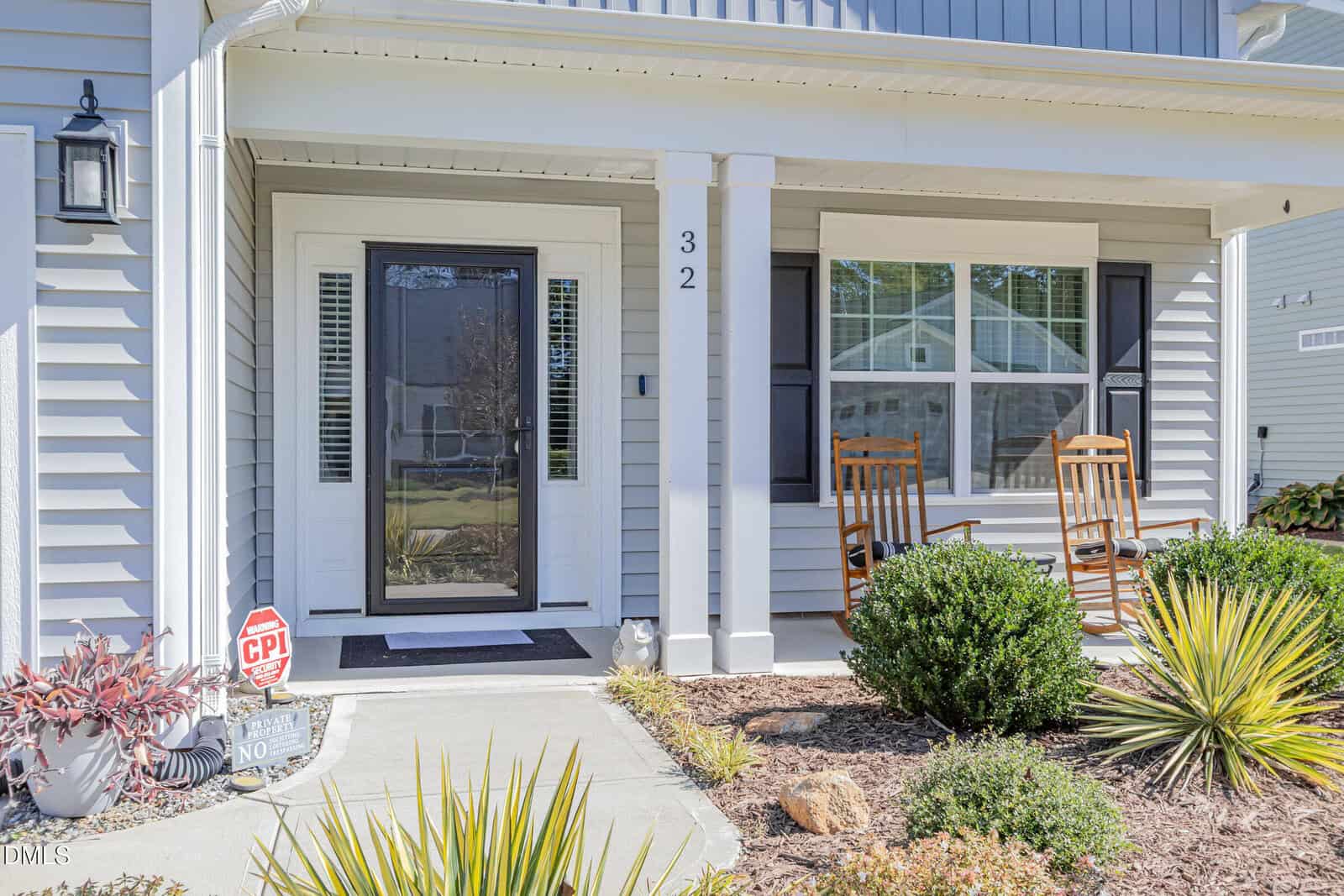 A front porch with two wooden rocking chairs, a glass storm door, house number 32, a CPI security sign, shrubs, and landscaped plants. The house exterior is light gray with white trim.