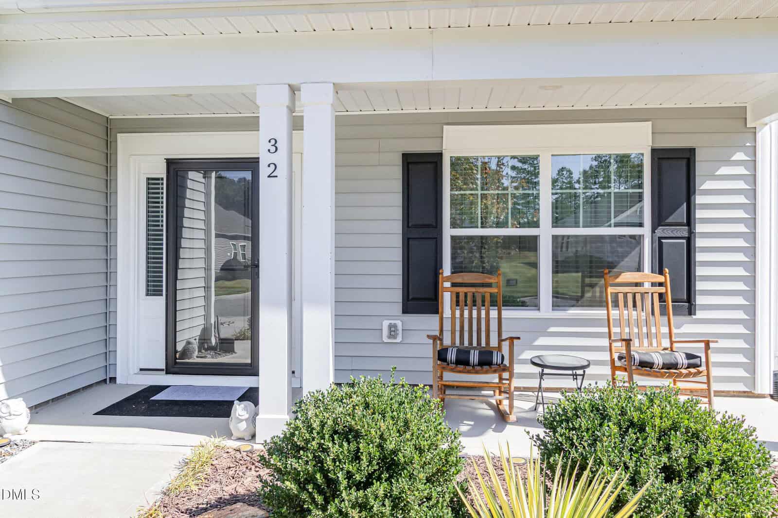 A covered front porch with two wooden rocking chairs, a small round table, and black shutters. The house has light gray siding, white trim, and the number 32 is displayed on a porch post. Shrubs line the edge of the porch.