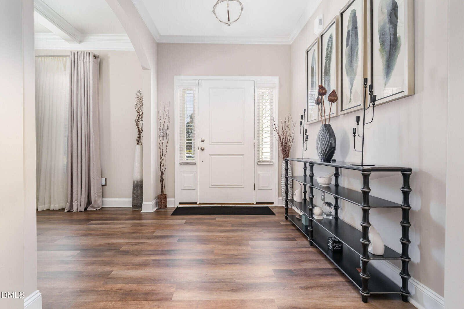 A modern entryway with wood flooring, a white front door, two narrow windows, contemporary decor on a black console table, tall vases, and framed artwork on a beige wall. Light fills the space.
