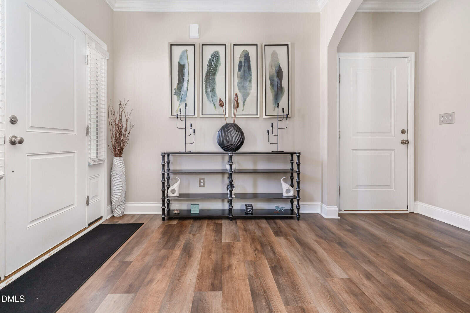 A tidy entryway with wood flooring, a black shelving unit holding decor items, and four feather artwork pieces above it. Two white doors and a tall vase with dry branches are also visible.