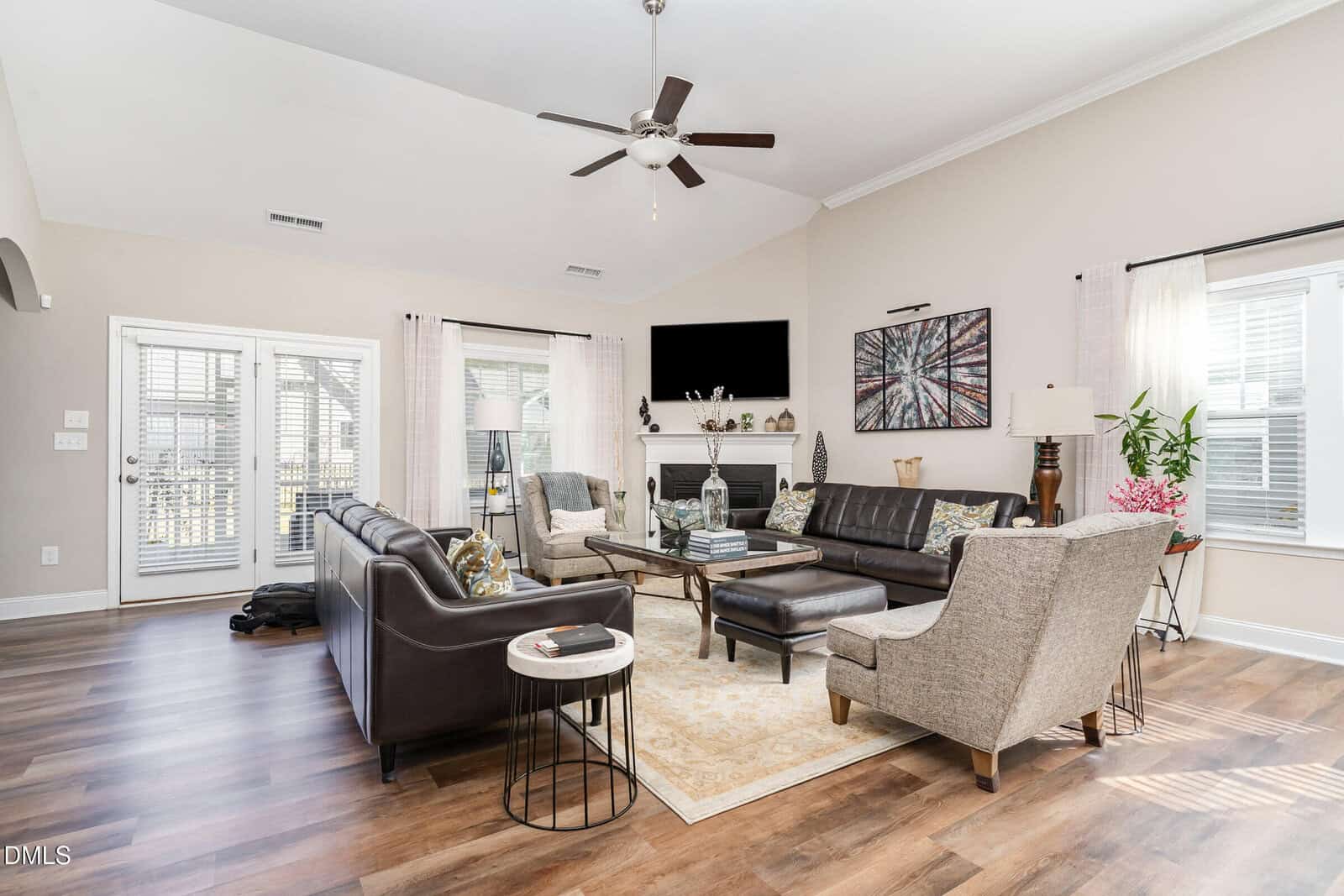 A bright living room with wood floors, a ceiling fan, and neutral walls. It features a brown leather sofa, two beige armchairs, a fireplace, a TV, large windows with light curtains, and various decor elements.