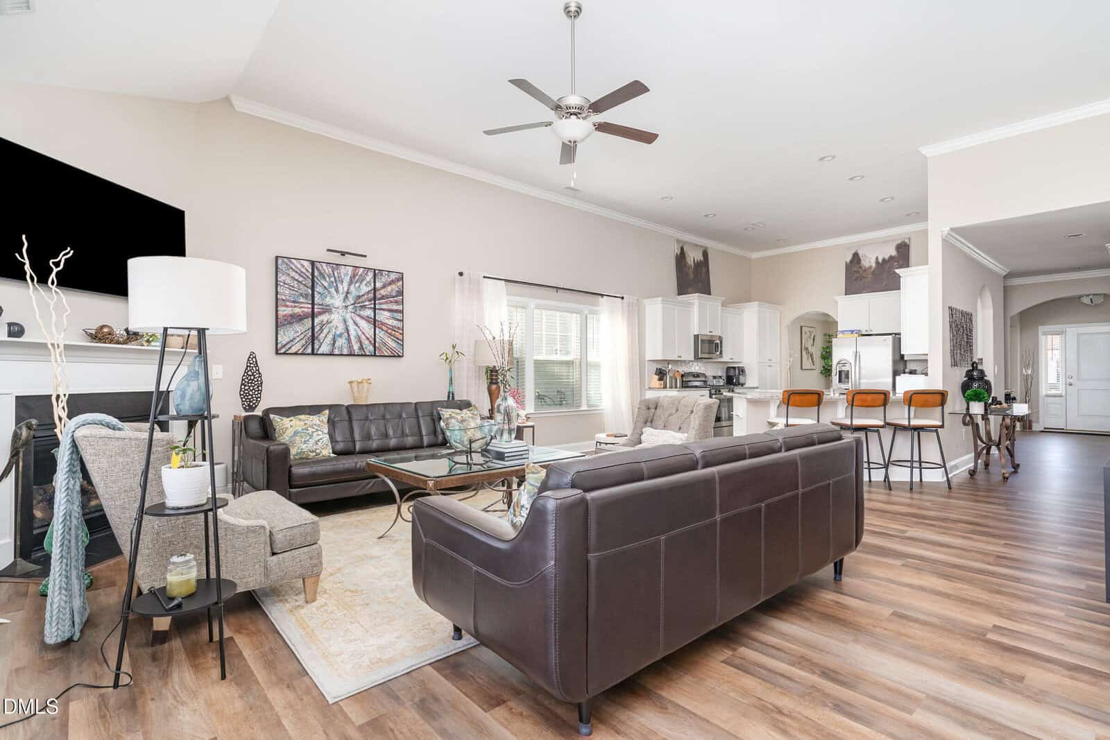 Open-concept living space with dark leather couches, a coffee table, and accent chairs. The kitchen features white cabinets, stainless steel appliances, and a breakfast bar with wooden stools. There is wood flooring throughout.